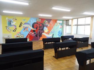 ピアノ室の写真。電子ピアノが教室内に並んでいる。教室の後ろには美術工芸部による壁画が掲載されている。