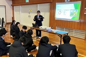 スクリーンの前で研究成果を発表する3年生1名と、説明を聞く後輩たちの写真