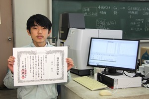 環境化学科2年生の滝本さんの写真、手に賞状を持ち、横には学校の分析機器がある