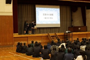 代表グループが体育館の舞台でモニターを使って発表している写真