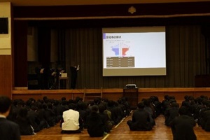 代表グループが体育館の舞台でモニターを使って発表している写真