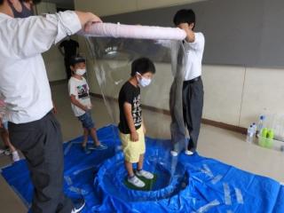 二人の生徒が大きなシャボン玉の輪を使い、小学生をシャボン玉で包んでいる。