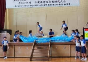 体育館の舞台でサーフィンデモンストレーションを披露する児童たちの写真