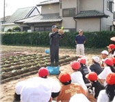 畑の前で説明をする地域の方と、説明を聞く児童たちの写真