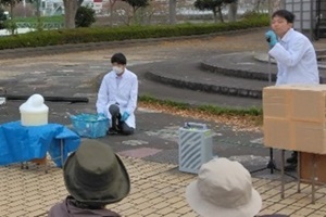 白衣姿で実験を披露する生徒2名と、それを見守る観客の写真