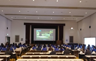 ホールに集まり、事前学習として見学する企業の説明を受けている中学生たちの写真